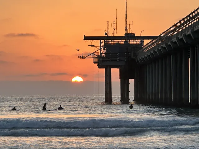 The setting sun beside a pier with people in the surf below.